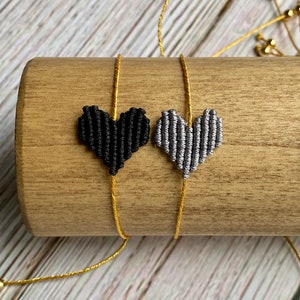 Puede incluir: Dos pulseras con dijes en forma de coraz&oacute;n hechos de cord&oacute;n trenzado. Un coraz&oacute;n es negro y el otro es gris plateado. Cada dije est&aacute; centrado en un cord&oacute;n fino de color dorado met&aacute;lico con cuentas deslizantes ajustables en los extremos.