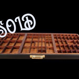May include: A vintage wooden printer's tray with numerous small compartments. The tray is rectangular, with a dark brown frame and a lighter wood interior. A white sign that says "SOLD" is above the tray.