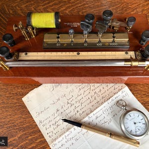 May include: Antique electrical resistance box with brass knobs and a wooden base. A yellow coil is visible, along with a ruler and various terminals. A handwritten letter, a pocket watch, and a pen are also present.