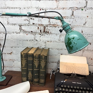 May include: An adjustable, vintage-style desk lamp with a teal green finish and a black cord. The lamp is positioned over a vintage typewriter, old books, and a sheet of paper, set against a white brick wall.