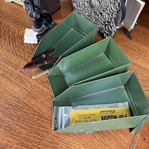 May include: Three green metal trays stacked on a wooden surface. The top tray contains a yellow package with the text "Felt-Tip Pen with Valve Control" and "Product of  Gablebrook".