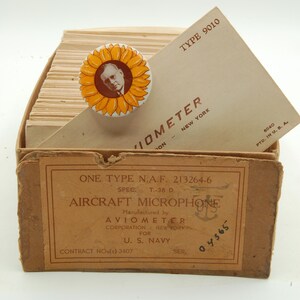 May include: A vintage box labeled "ONE TYPE N.A.F. 213264-6 AIRCRAFT MICROPHONE" with text from the manufacturer, Aviometer Corporation, for the U.S. Navy. A sunflower pin with a portrait rests on top, along with a tan envelope.