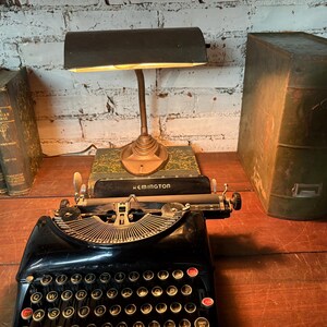 May include: A vintage black Remington typewriter with a gold-colored metal trim, a desk lamp, and several antique books. The lamp has a dark green shade and is positioned above the typewriter. The books are bound in dark green and gold.