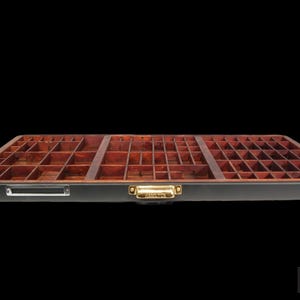 May include: A rectangular wooden printer's type drawer featuring numerous small compartments of varying sizes. The tray has a dark brown finish, a black outer frame, a brass-colored metal handle labeled Hamilton, and a silver-toned label holder.
