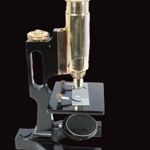 May include: An antique microscope with a black base and a brass-colored barrel. The microscope has a stage for slides and a focusing knob. The image is set against a black background.