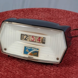 May include: Black and white flip clock with a white face and black numbers. The clock displays the time 12:54. The clock has a small label that says "First Orbit" with a rocket ship graphic.