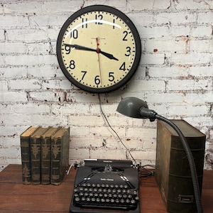 May include: A vintage-style wall clock with a black frame and a cream-colored face hangs above a desk. Below, a black typewriter, books, and a desk lamp are arranged on a wooden surface against a white brick wall.