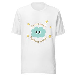 May include: White t-shirt featuring a teal cloud with a smiling face and the text "Current mood: Patching pimples" in brown. Yellow stars surround the cloud. A casual, humorous design.