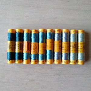 Op de afbeelding: Een verzameling van tien spoelen garen in verschillende tinten blauw en turquoise. De spoelen zijn verpakt in geel papier met zwarte tekst die "METRI" en "OCC" luidt.