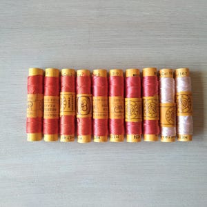 Può includere: Una collezione di 10 bobine di filo, 9 sono rosse e 1 è rosa. Le bobine sono avvolte in carta gialla con testo nero che recita "SUPER QUALITÀ" e "MADE IN ITALY".