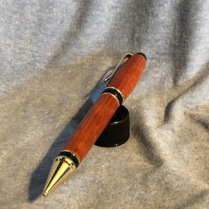 May include: A wooden pen with gold accents and black trim. The pen has a dark red wood grain and a gold clip.