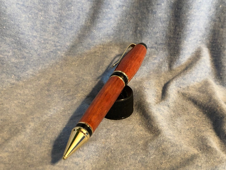 May include: A wooden pen with gold accents and black trim. The pen has a dark red wood grain and a gold clip.