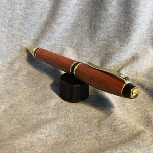 May include: A wooden pen with gold accents and black trim. The pen is resting on a black cap.