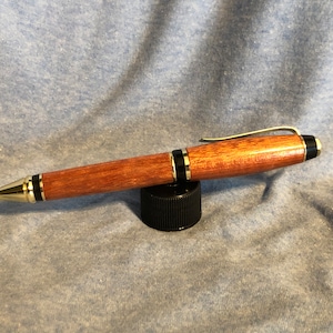 May include: A wooden pen with a gold and black trim. The pen is made of a reddish-brown wood and has a clip on the top.