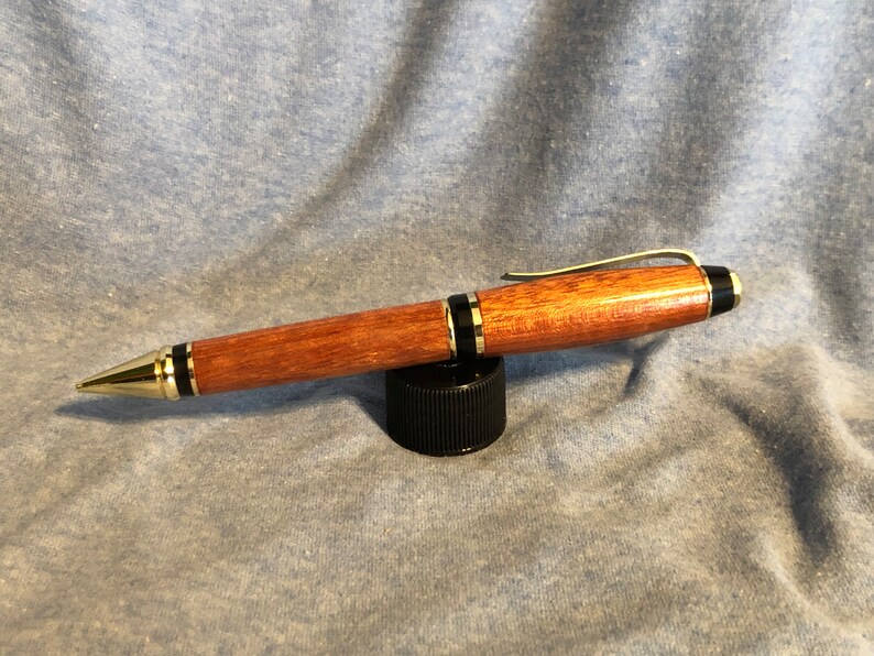 May include: A wooden pen with a gold and black trim. The pen is made of a reddish-brown wood and has a clip on the top.
