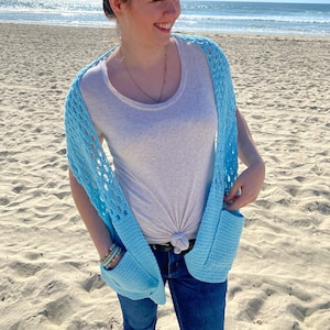 May include: A woman wearing a light blue crochet vest with pockets over a white t-shirt and blue jeans. She is standing on a sandy beach with the ocean in the background.