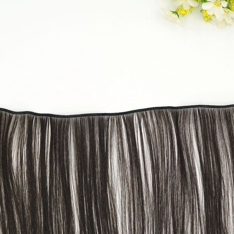 May include: A weft of dark brown and white hair extensions, with a black seam at the top. The hair has a natural, straight texture. The background is white, with a few yellow and white flowers in the upper right corner.