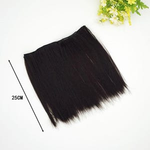 May include: A dark brown hair extension piece, approximately 25cm long, lies on a white surface. The hair appears straight and sleek, with a natural shine. A small bouquet of yellow and white flowers is in the background.