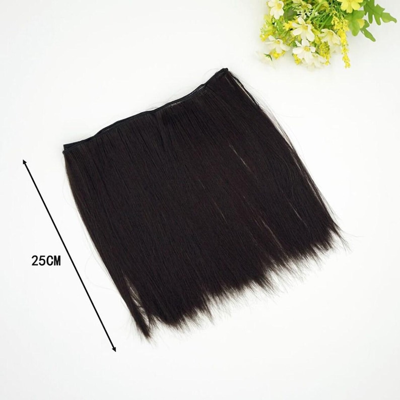May include: A dark brown hair extension piece, approximately 25cm long, lies on a white surface. The hair appears straight and sleek, with a natural shine. A small bouquet of yellow and white flowers is in the background.