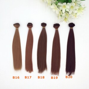 May include: Five bundles of hair extensions in varying shades of brown, from light to dark. Each bundle is tied at the top with a brown band. The labels B16, B17, B18, B19, and B20 are printed below each bundle. White flowers are in the background.