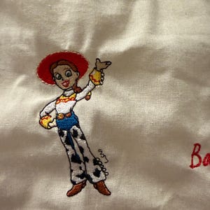 K&ouml;nnte beinhalten: Eine wei&szlig;e Stoffoberfl&auml;che mit einer gestickten Figur mit roten Haaren, einem roten Hut, einem wei&szlig;en Hemd mit gelben Akzenten, einer blauen Hose und einer wei&szlig;en Hose mit schwarzen Kuhflecken. Die Figur winkt und die roten Buchstaben Ba sind sichtbar.