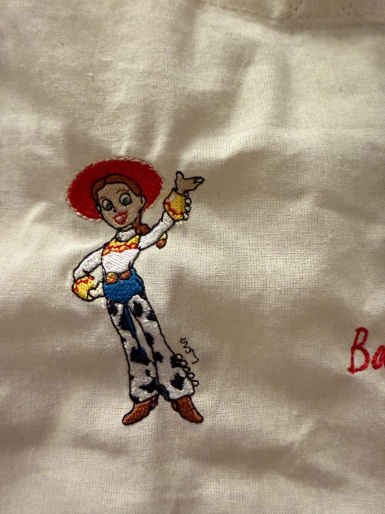 K&ouml;nnte beinhalten: Eine wei&szlig;e Stoffoberfl&auml;che mit einer gestickten Figur mit roten Haaren, einem roten Hut, einem wei&szlig;en Hemd mit gelben Akzenten, einer blauen Hose und einer wei&szlig;en Hose mit schwarzen Kuhflecken. Die Figur winkt und die roten Buchstaben Ba sind sichtbar.