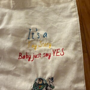 K&ouml;nnte beinhalten: Eine cremefarbene Stofftasche mit gesticktem Text, der It's a Toy Story Baby just say YES in blauem, gelbem und rotem Garn zeigt. Unter dem Text befindet sich ein gestickter Aufn&auml;her der Figur Buzz Lightyear in seinem Raumanzug.