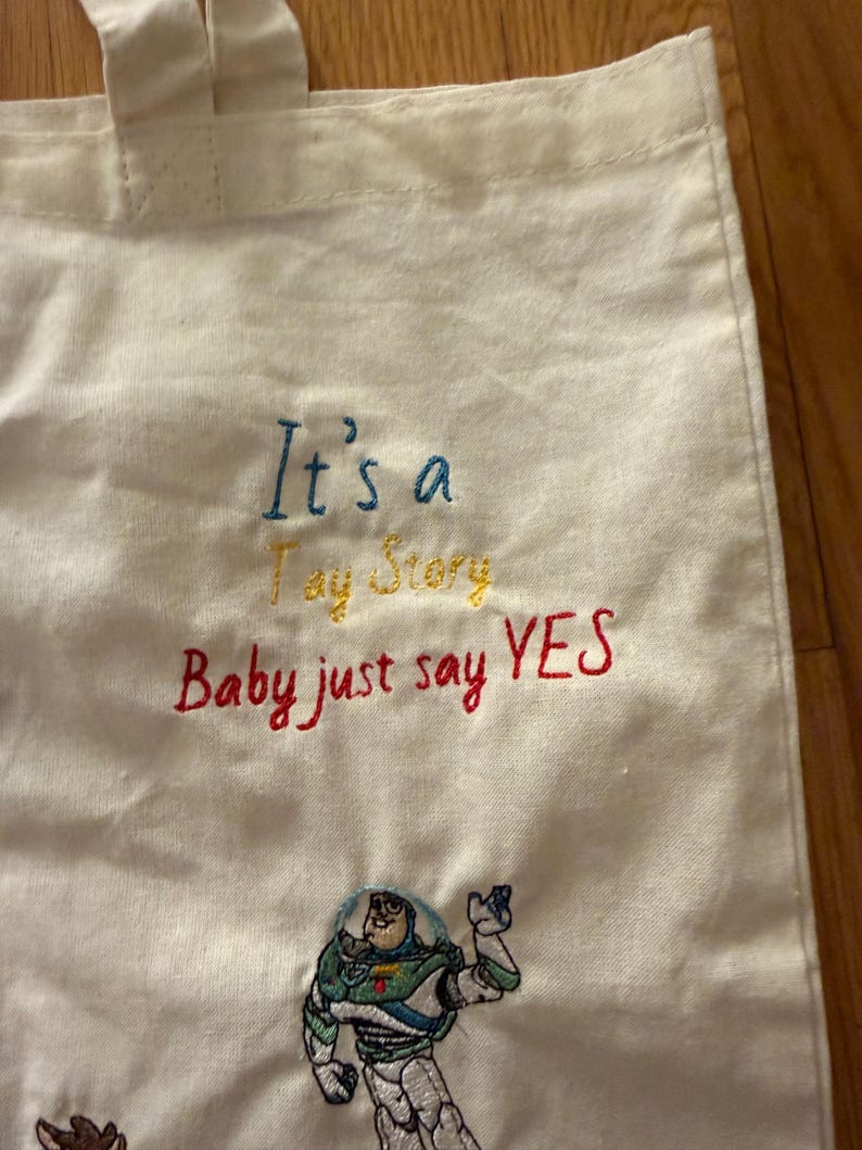 K&ouml;nnte beinhalten: Eine cremefarbene Stofftasche mit gesticktem Text, der It's a Toy Story Baby just say YES in blauem, gelbem und rotem Garn zeigt. Unter dem Text befindet sich ein gestickter Aufn&auml;her der Figur Buzz Lightyear in seinem Raumanzug.
