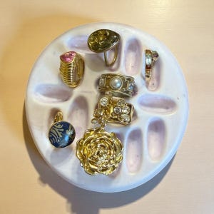 May include: A collection of gold-toned rings and a pendant displayed on a white and pink ceramic ring holder. The rings feature various designs, including a pink gemstone, pearls, and clear crystals. The pendant is a dark blue with a gold design.
