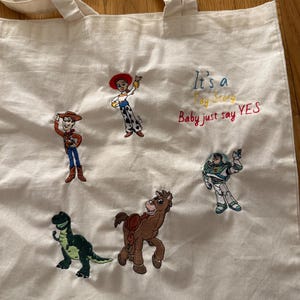K&ouml;nnte beinhalten: Eine cremefarbene Stofftasche mit gestickten Toy Story-Figuren, darunter Woody, Jessie, Buzz Lightyear, Rex und Bullseye. Der Text "It's a Toy Story Baby just say YES" ist in blauem, gelbem und rotem Garn auf die Tasche gestickt.