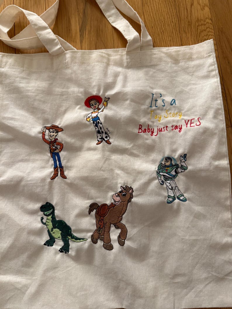 K&ouml;nnte beinhalten: Eine cremefarbene Stofftasche mit gestickten Toy Story-Figuren, darunter Woody, Jessie, Buzz Lightyear, Rex und Bullseye. Der Text "It's a Toy Story Baby just say YES" ist in blauem, gelbem und rotem Garn auf die Tasche gestickt.