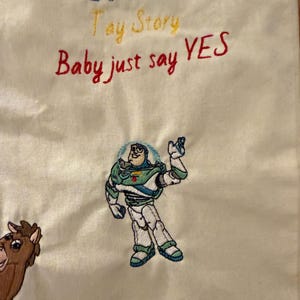 K&ouml;nnte beinhalten: Ein cremefarbener Stoff mit gesticktem Text, der It's a Toy Story Baby just say YES lautet. Unter dem Text befindet sich eine gestickte Figur eines Weltraum-Ranger-Charakters in einem wei&szlig;-gr&uuml;n-violetten Anzug neben einem braunen Pferdecharakter.