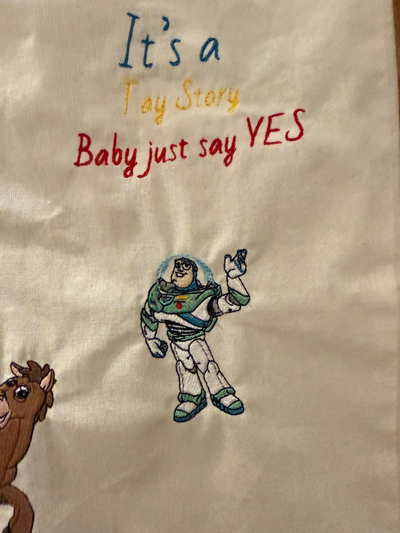 K&ouml;nnte beinhalten: Ein cremefarbener Stoff mit gesticktem Text, der It's a Toy Story Baby just say YES lautet. Unter dem Text befindet sich eine gestickte Figur eines Weltraum-Ranger-Charakters in einem wei&szlig;-gr&uuml;n-violetten Anzug neben einem braunen Pferdecharakter.