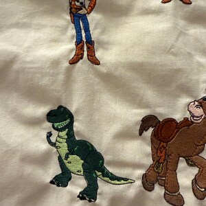 K&ouml;nnte beinhalten: Ein cremefarbener Stoff mit gestickten Figuren aus der Toy Story-Reihe, darunter ein Cowboy in gelbem Hemd und blauer Jeans, ein gr&uuml;ner Dinosaurier und ein braunes Pferd mit Sattel, die alle in einem wiederkehrenden Muster angeordnet sind.