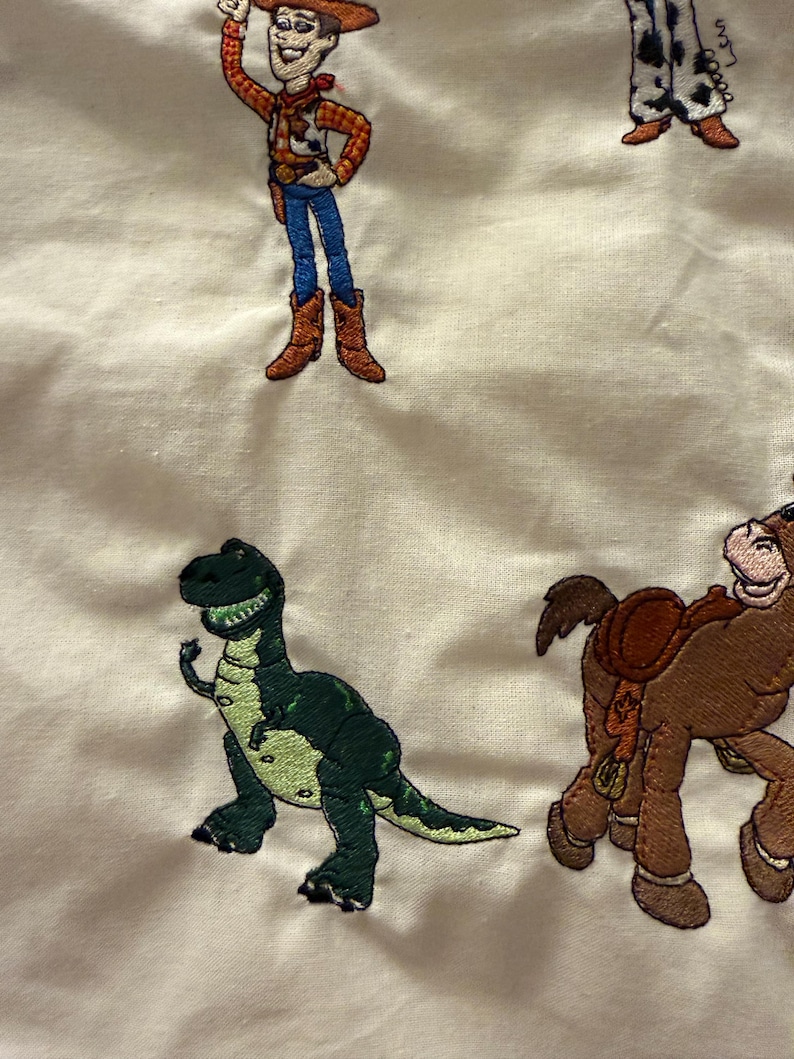 K&ouml;nnte beinhalten: Ein cremefarbener Stoff mit gestickten Figuren aus der Toy Story-Reihe, darunter ein Cowboy in gelbem Hemd und blauer Jeans, ein gr&uuml;ner Dinosaurier und ein braunes Pferd mit Sattel, die alle in einem wiederkehrenden Muster angeordnet sind.