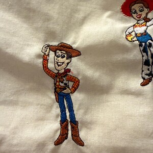 K&ouml;nnte beinhalten: Ein cremefarbener Stoff mit zwei gestickten Figuren. Links tr&auml;gt eine Cowboy-Figur einen braunen Hut, ein gelb-rotes kariertes Hemd, eine blaue Hose und braune Stiefel. Rechts tr&auml;gt eine Cowgirl-Figur einen roten Hut und ein wei&szlig;es Hemd.