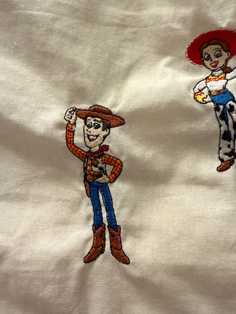 K&ouml;nnte beinhalten: Ein cremefarbener Stoff mit zwei gestickten Figuren. Links tr&auml;gt eine Cowboy-Figur einen braunen Hut, ein gelb-rotes kariertes Hemd, eine blaue Hose und braune Stiefel. Rechts tr&auml;gt eine Cowgirl-Figur einen roten Hut und ein wei&szlig;es Hemd.