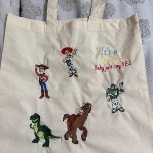 K&ouml;nnte beinhalten: Eine cremefarbene Stofftasche mit gestickten Toy Story-Figuren, darunter Woody, Jessie, Buzz Lightyear, Rex und Bullseye. Der Text "It's a Toy Story Baby just say YES" ist in blauem, gelbem und rotem Garn auf die Tasche gestickt.