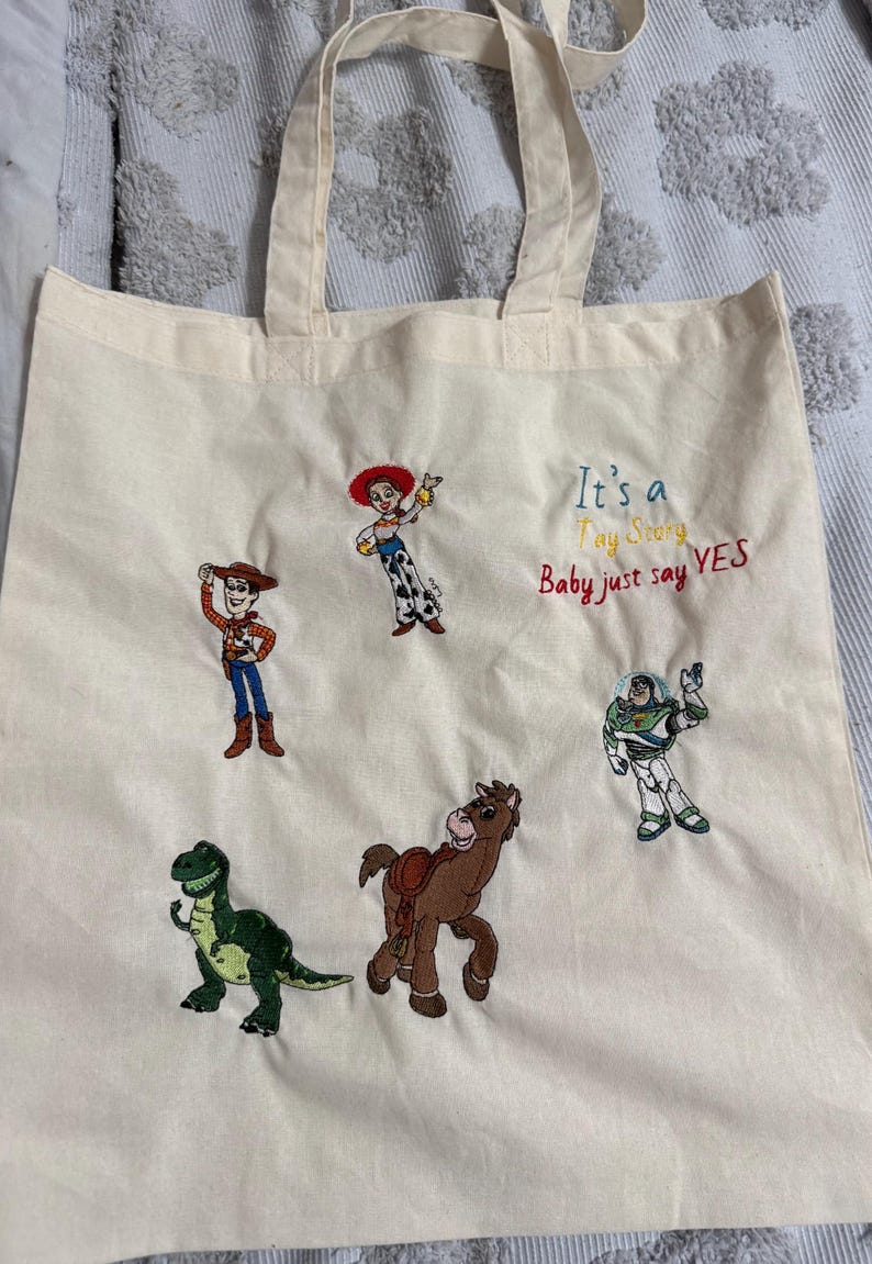 K&ouml;nnte beinhalten: Eine cremefarbene Stofftasche mit gestickten Toy Story-Figuren, darunter Woody, Jessie, Buzz Lightyear, Rex und Bullseye. Der Text "It's a Toy Story Baby just say YES" ist in blauem, gelbem und rotem Garn auf die Tasche gestickt.