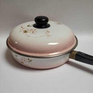 Puede incluir: Una sartén redonda de metal con tapa a juego, que presenta un degradado de color blanco y rosa suave. El exterior y la tapa están decorados con delicados motivos florales en tonos apagados. Incluye un mango negro y un pomo negro en la tapa.