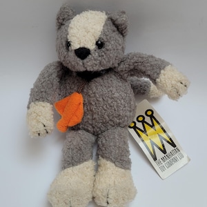 May include: A plush cat toy with gray and cream-colored textured fur, black eyes, and a black nose. The cat wears a dark collar with an orange leaf-shaped charm. A white tag attached to the side features a yellow crown logo and The Manhattan Toy Company Ltd.