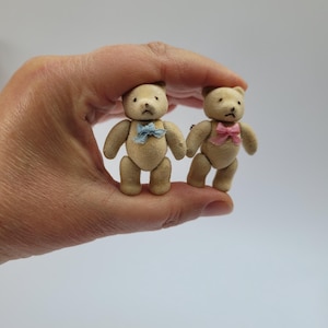 Könnte beinhalten: Zwei Miniatur-Teddybären mit beigefarbenem Fell werden in einer Hand gehalten. Einer der Bären hat eine blaue Fliege, der andere eine rosa Fliege. Die Bären haben kleine schwarze Augen und stehen aufrecht.