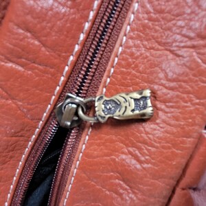 May include: A textured rust-colored leather bag featuring a brown zipper with white contrast stitching. The zipper pull is a small, metallic, antique-brass colored charm shaped like a dog, attached to the zipper slider by a circular metal ring.