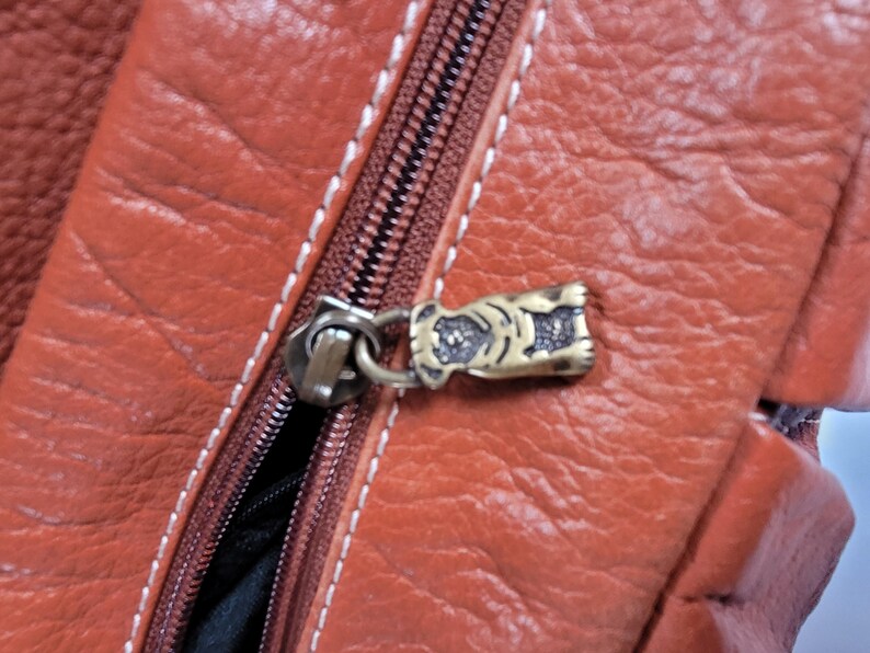 May include: A textured rust-colored leather bag featuring a brown zipper with white contrast stitching. The zipper pull is a small, metallic, antique-brass colored charm shaped like a dog, attached to the zipper slider by a circular metal ring.