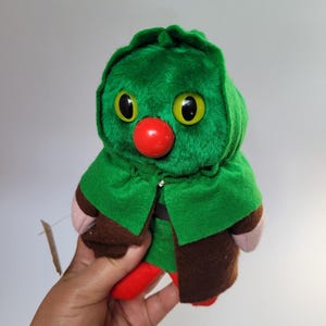 May include: A plush toy featuring a green hooded figure with large yellow eyes, a red nose, and brown and red clothing. The toy is held in a hand against a plain background. The toy has a tag attached.