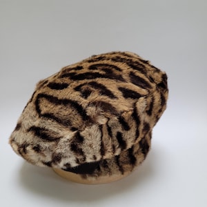 Puede incluir: Gorra de estilo newsboy con estampado marrón y negro. El sombrero tiene una corona redondeada y una visera corta y rígida. La tela tiene una textura de piel sintética con manchas marrones oscuras sobre un fondo marrón claro. La gorra está sobre un fondo blanco.