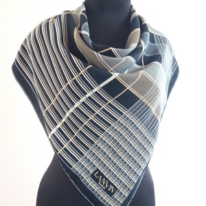 Könnte beinhalten: Ein quadratisches Tuch mit dunkelblauem Hintergrund und einem geometrischen Muster aus sich kreuzenden weißen, grauen und beigen Linien. Das Tuch zeigt den Schriftzug LANVIN PARIS auf einem rechteckigen Etikett nahe der unteren Ecke.