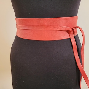 Può includere: Un'ampia cintura in stile obi in pelle color rosso corallo con una superficie testurizzata. La cintura si avvolge attorno alla vita e si fissa con un lungo e sottile laccio in pelle annodato sul lato destro, creando due estremità pendenti.
