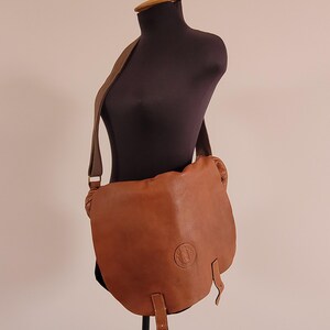 May include: A tan leather messenger bag with a wide shoulder strap and a large front flap. The bag features a circular embossed logo on the lower right side of the flap and two vertical leather straps hanging from the bottom edge of the flap.