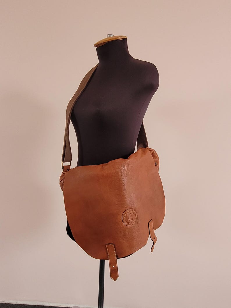 May include: A tan leather messenger bag with a wide shoulder strap and a large front flap. The bag features a circular embossed logo on the lower right side of the flap and two vertical leather straps hanging from the bottom edge of the flap.